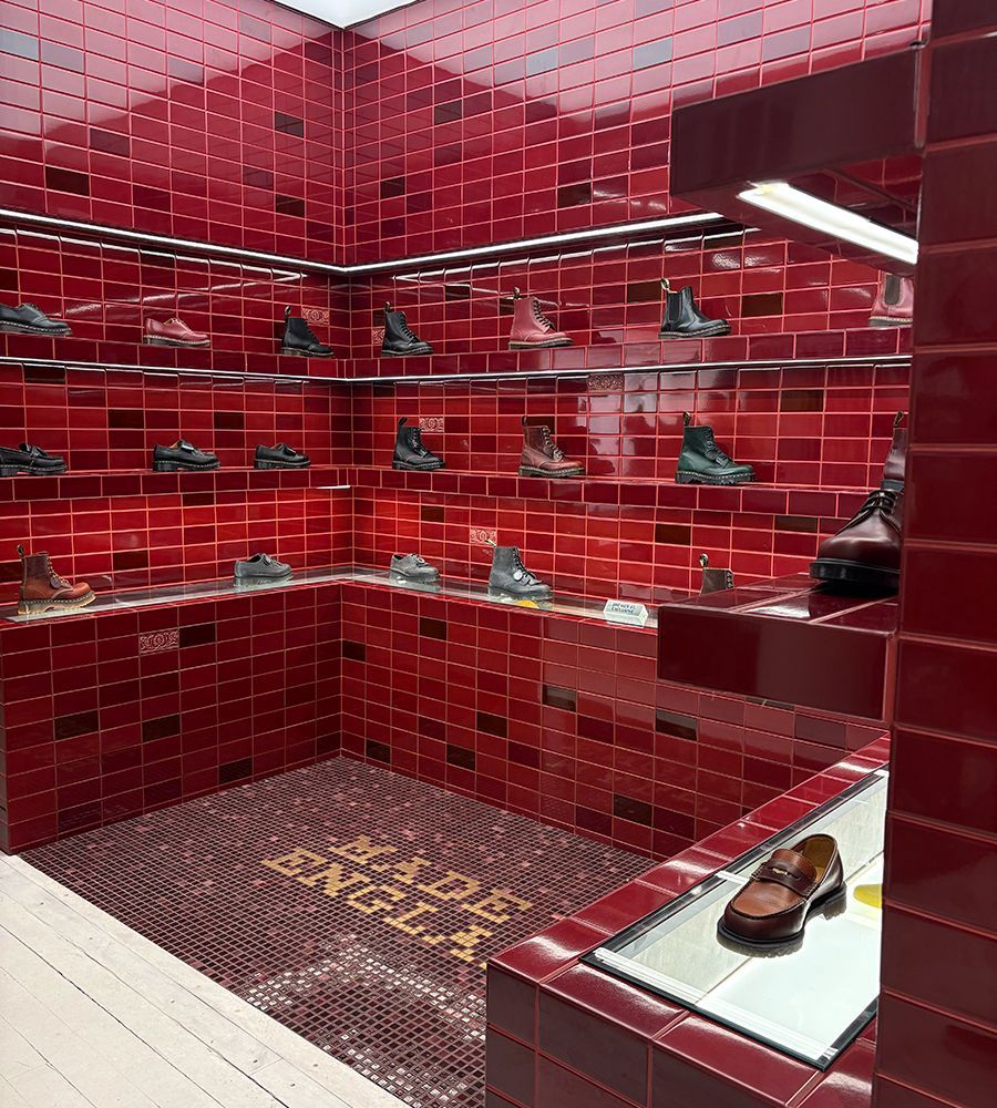 Display of various boots and shoes on multiple glass shelves in a corner room with glossy red tiled walls and floor, the floor tiles spelling out "DR. MARTENS ENGLAND."
