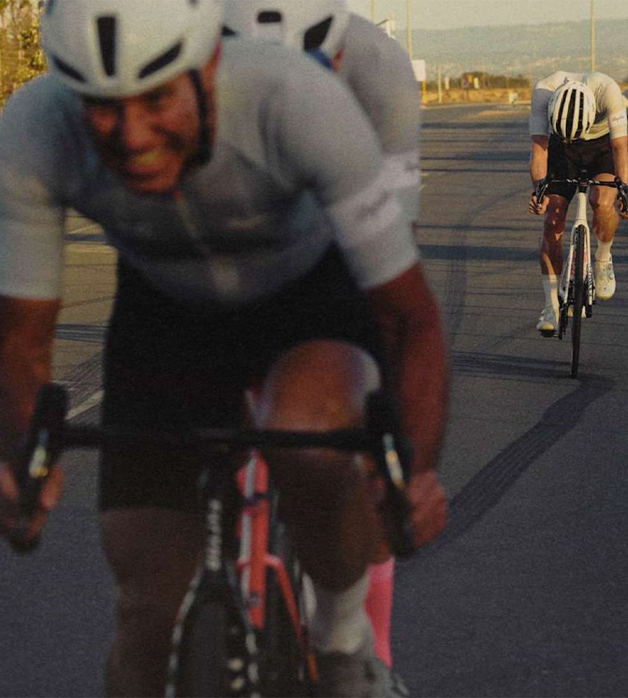 Two cyclists wearing white helmets and matching cycling outfits are riding on a road in a low stance, with the cyclist in the foreground slightly blurred and the cyclist in the background clearly visible. The road has visible tire marks and the setting appears to be during sunset or sunrise.