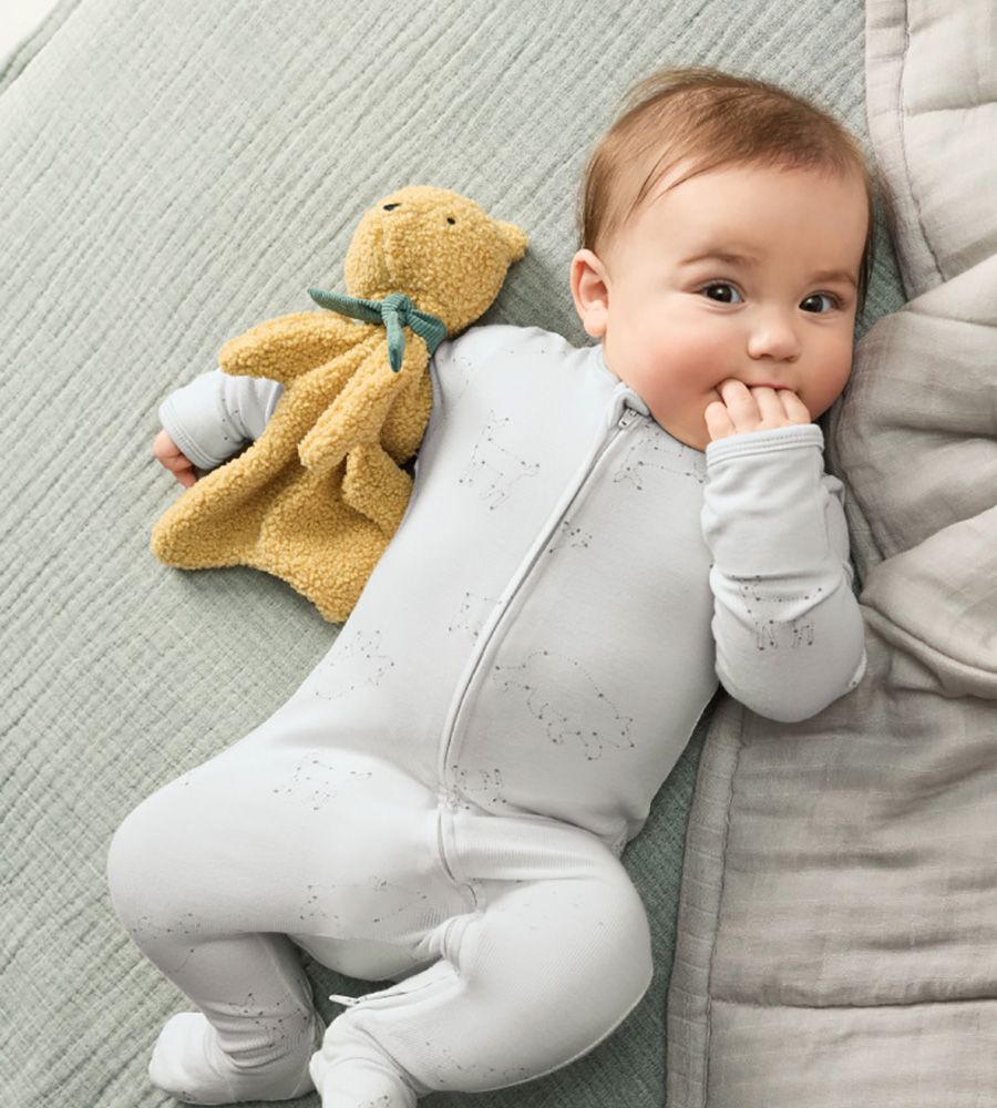 Baby in a light grey sleeper lying on a grey quilted blanket with a small tan plush bear blanket beside them, with fingers in their mouth.