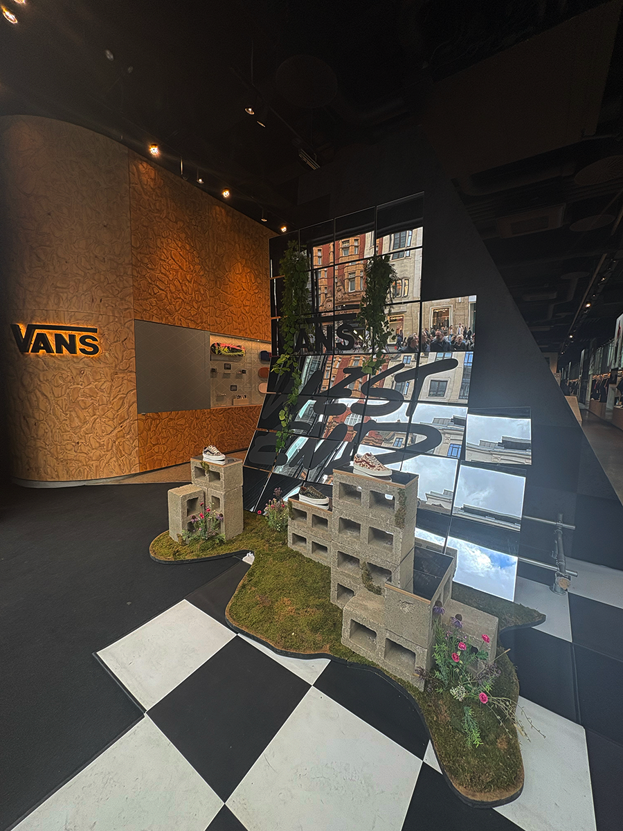 Retail display featuring Vans shoes placed on cinder blocks with grass and flower decor, set against a mirrored wall reflecting a street scene and black-and-white checkered flooring, inside a Vans store with wooden textured walls.